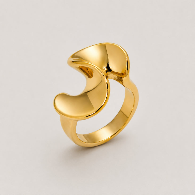A polished gold abstract ring with a sculptural, flowing design, photographed on a neutral light gray background.