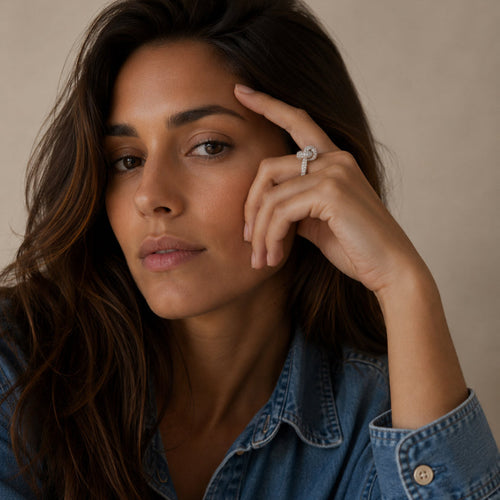 A close-up portrait of a woman with long dark brown hair wearing a blue denim shirt and a silver ring with a clear gemstone on her finger, set against a warm beige background.