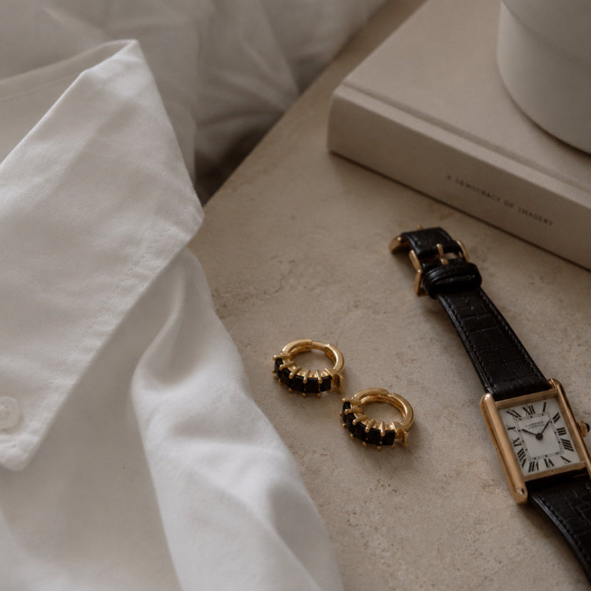 Two gold rings with black bead details are displayed on a beige stone surface next to a black leather strap watch and a white shirt, with a book and a white ceramic object in the background.