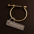 Gold-toned metal hoop earring with a sliding bar clasp, shown on a dark brown background with a branded hangtag reading Matriarcha.