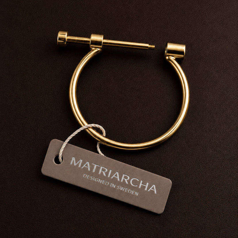 Gold-toned metal hoop earring with a sliding bar clasp, shown on a dark brown background with a branded hangtag reading Matriarcha.