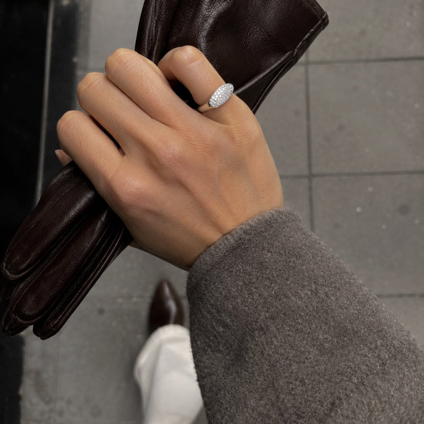 A close-up of a hand holding a pair of dark brown leather gloves while wearing a silver ring with a textured, diamond-like surface. The person is also wearing a gray knit sweater and white pants, standing on a gray tiled floor.