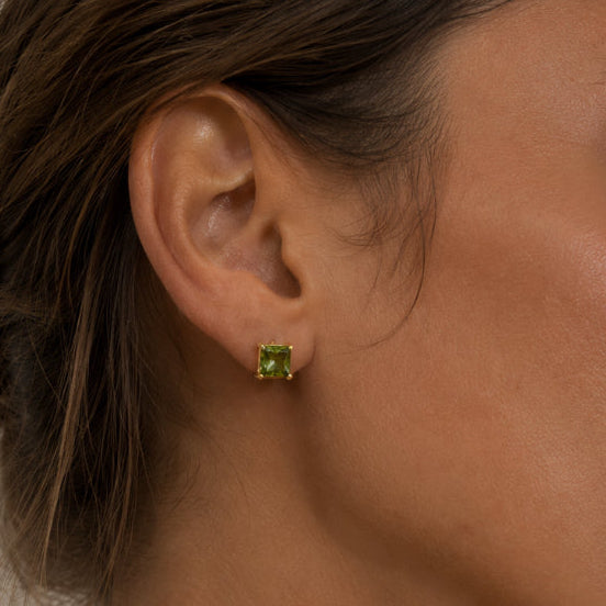 A close-up of a woman's ear wearing a small square green stud earring with a gold setting against a soft neutral background.