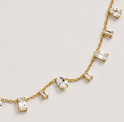 A gold chain necklace featuring multiple dangling clear gemstones in various shapes, including pear, baguette, round, and marquise cuts, displayed on a light gray background.