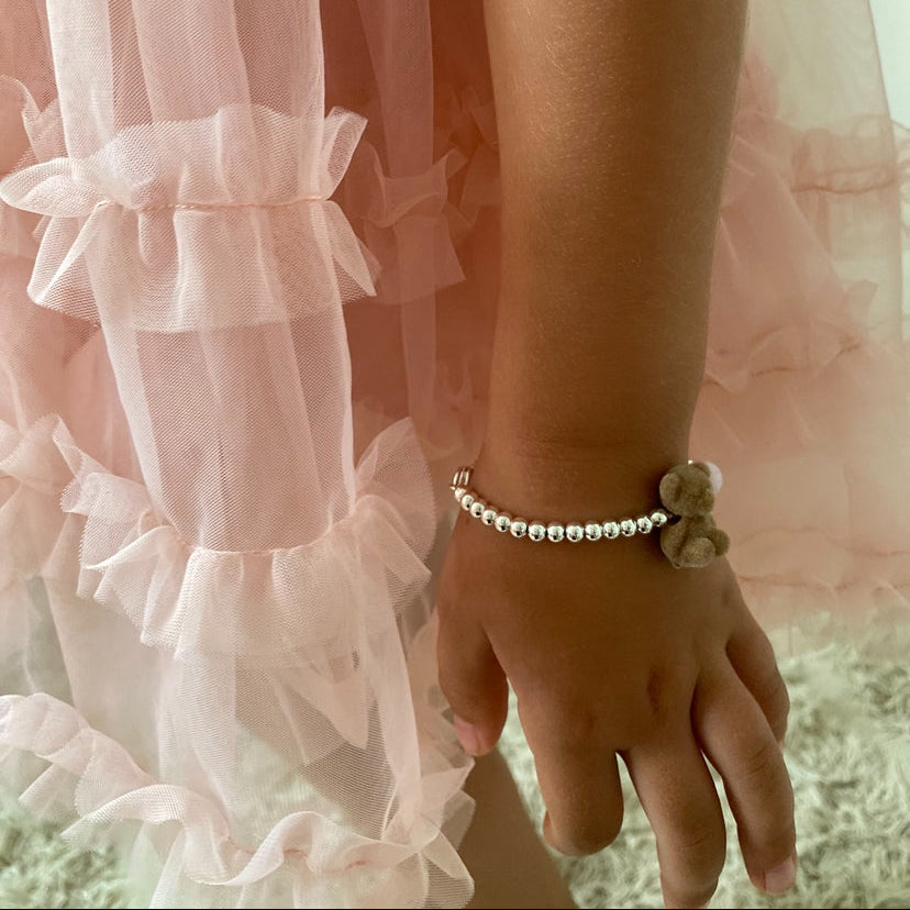 A close-up of a child's hand and wrist wearing a beaded bracelet with a small clover charm, next to a light pink ruffled tulle skirt.