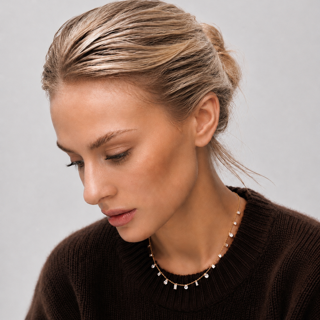 A close-up of a woman with blonde hair in a low bun wearing a delicate gold necklace with small dangling charms, paired with a dark brown knit sweater against a light gray background.