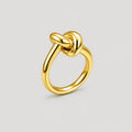 A polished yellow gold ring featuring a knotted design at the top, shown on a neutral light gray background.