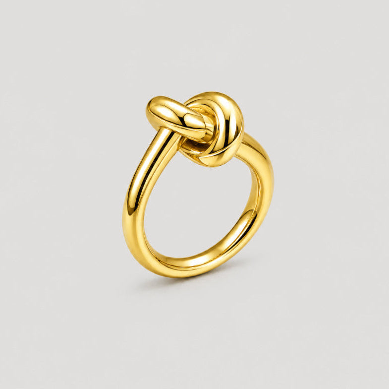A polished yellow gold ring featuring a knotted design at the top, shown on a neutral light gray background.