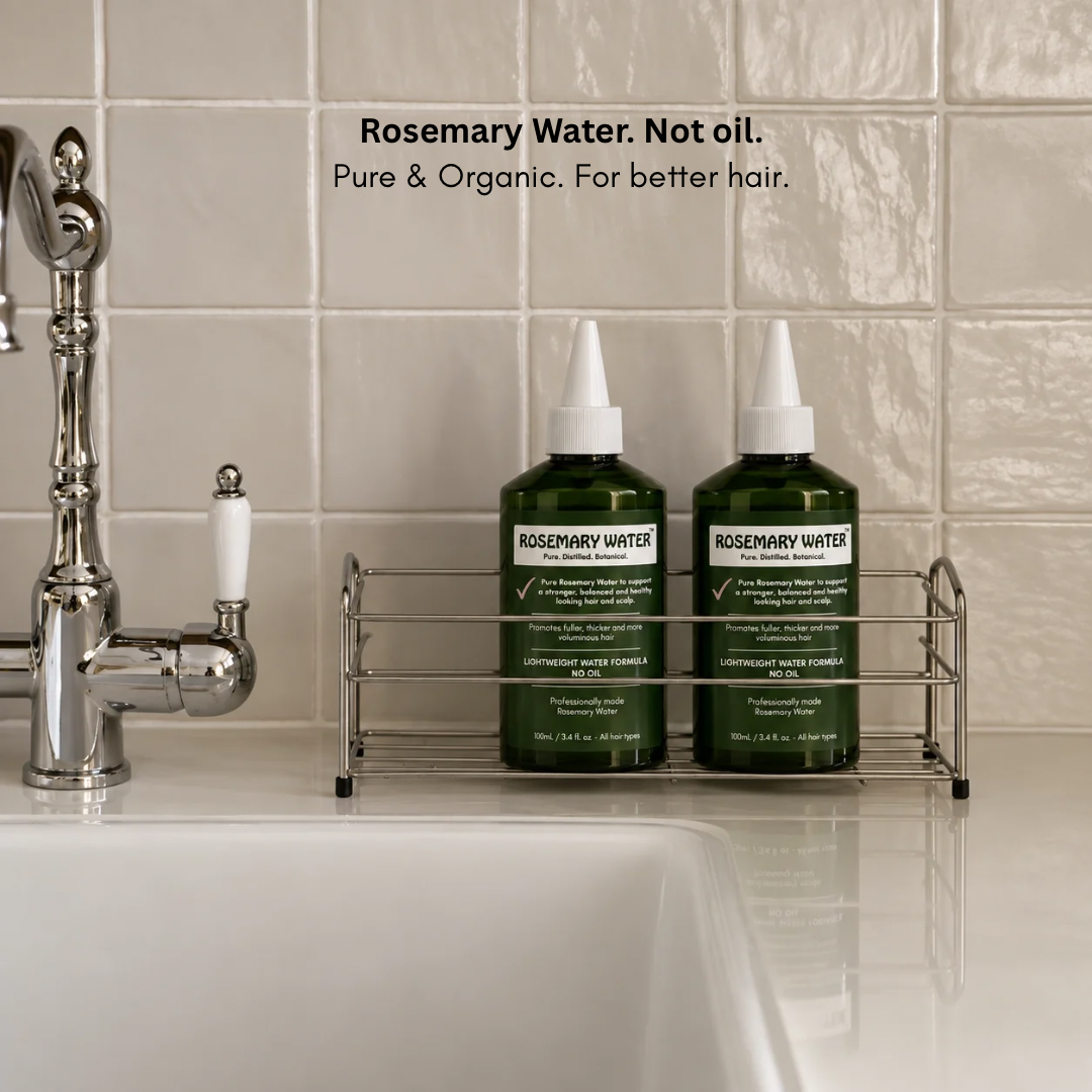 Two dark green Rosemary Water hair treatment bottles displayed on a chrome bathroom sink rack next to a faucet, with text overlay reading 'Rosemary Water. Not oil.