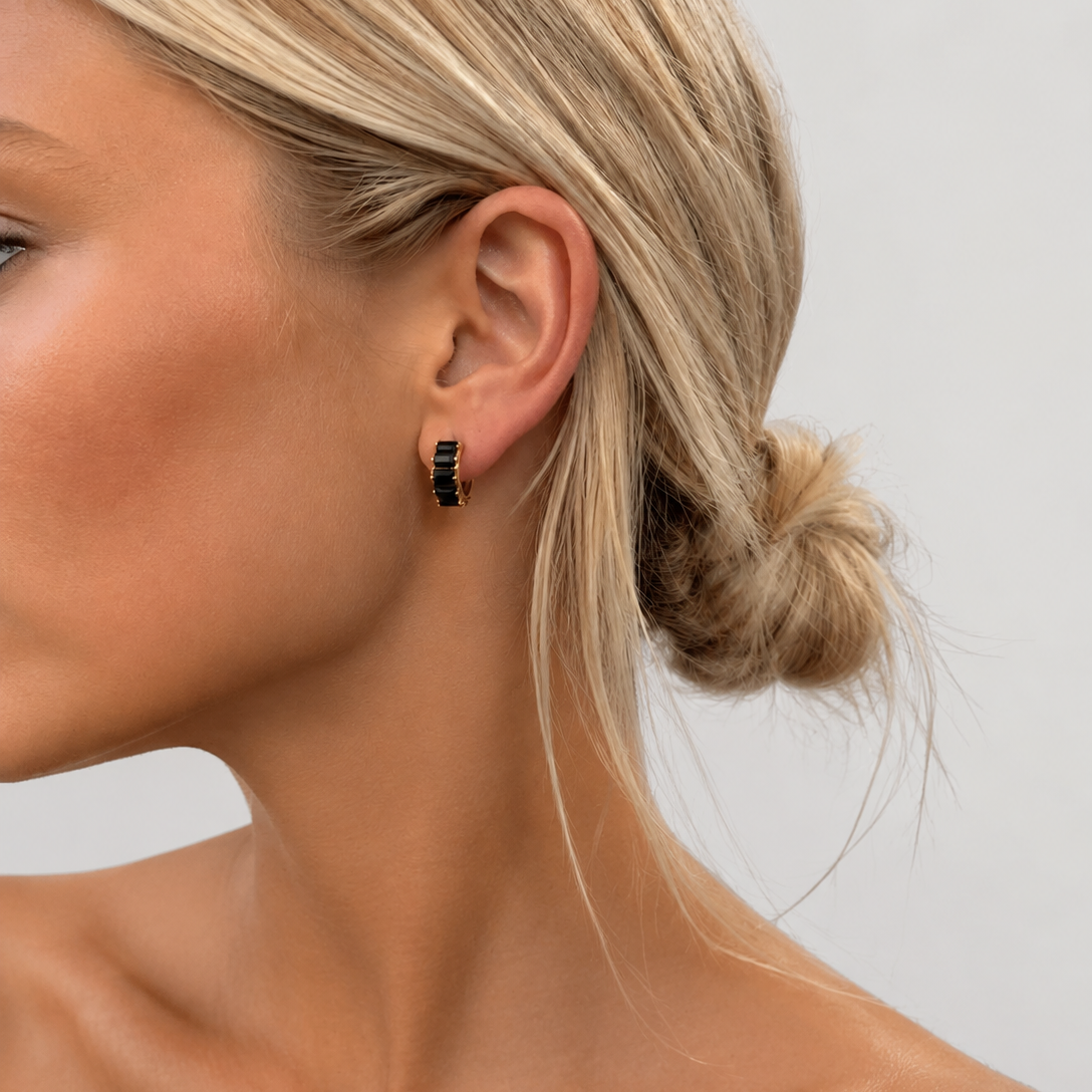 A close-up side profile of a woman with blonde hair in a low bun, wearing a small black and gold geometric hoop earring against a light gray background.