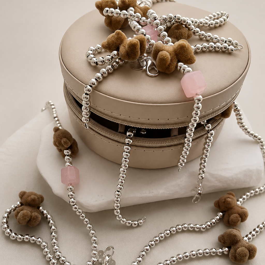 A beige round jewelry box with a zipper, styled with silver beaded necklaces featuring small brown teddy bear charms and pink stone accents, set against a warm beige background.