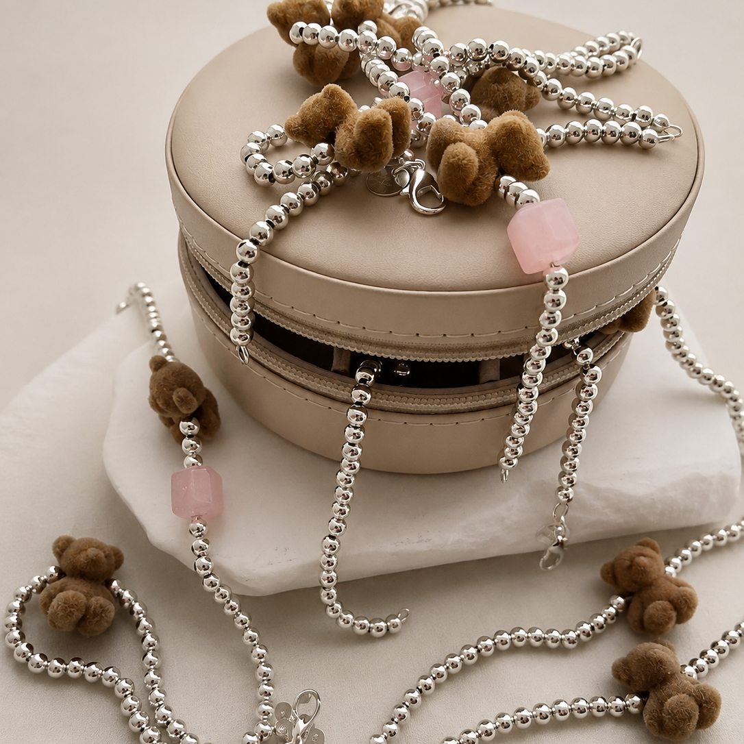 A beige round jewelry box with a zipper, styled with silver beaded necklaces featuring small brown teddy bear charms and pink stone accents, set against a warm beige background.