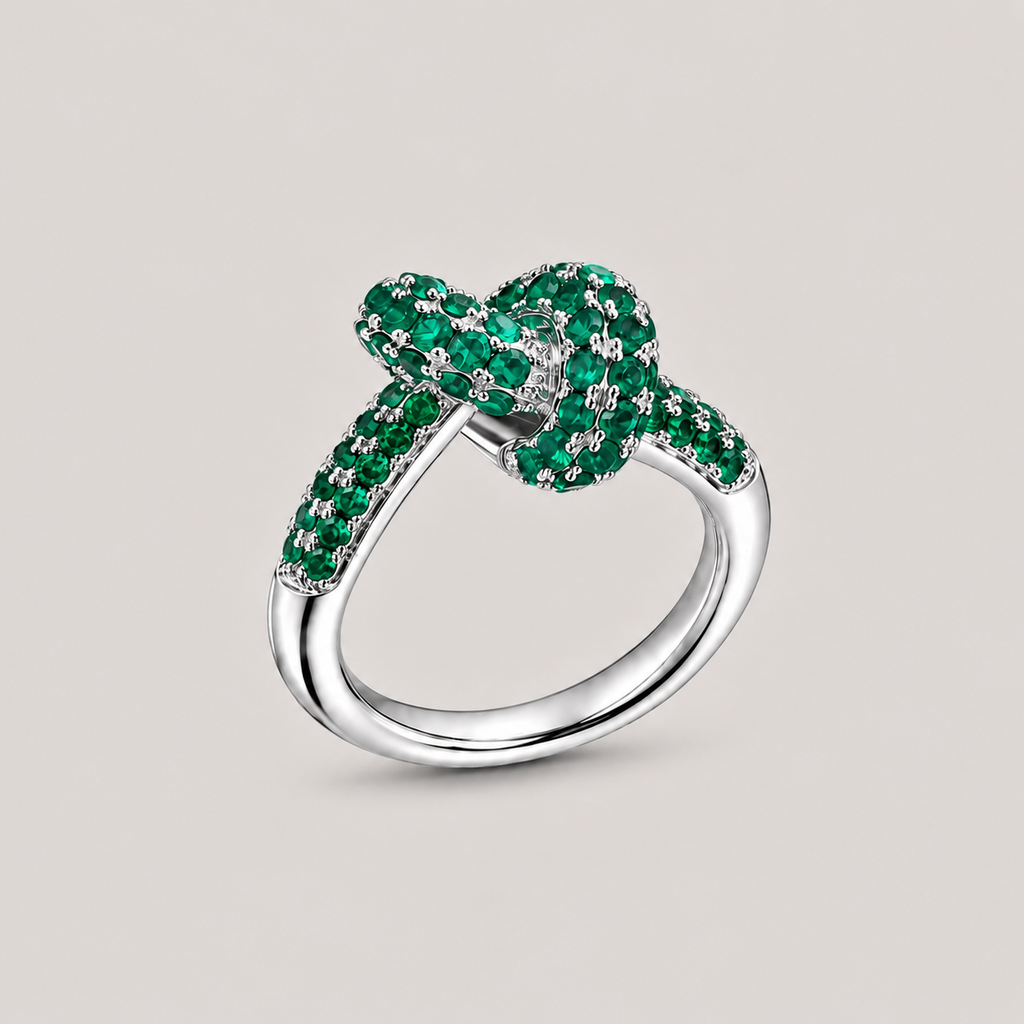 A silver ring featuring a knotted bow design paved with vibrant green emeralds, set against a solid light gray background.