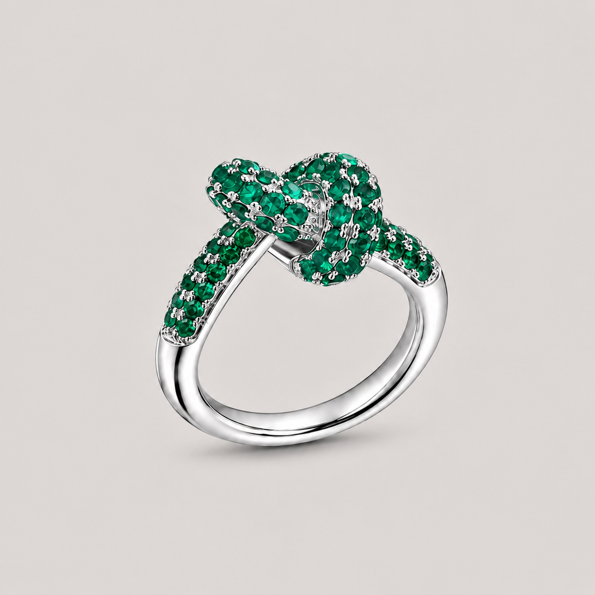 A silver ring featuring a knotted bow design paved with vibrant green emeralds, set against a solid light gray background.