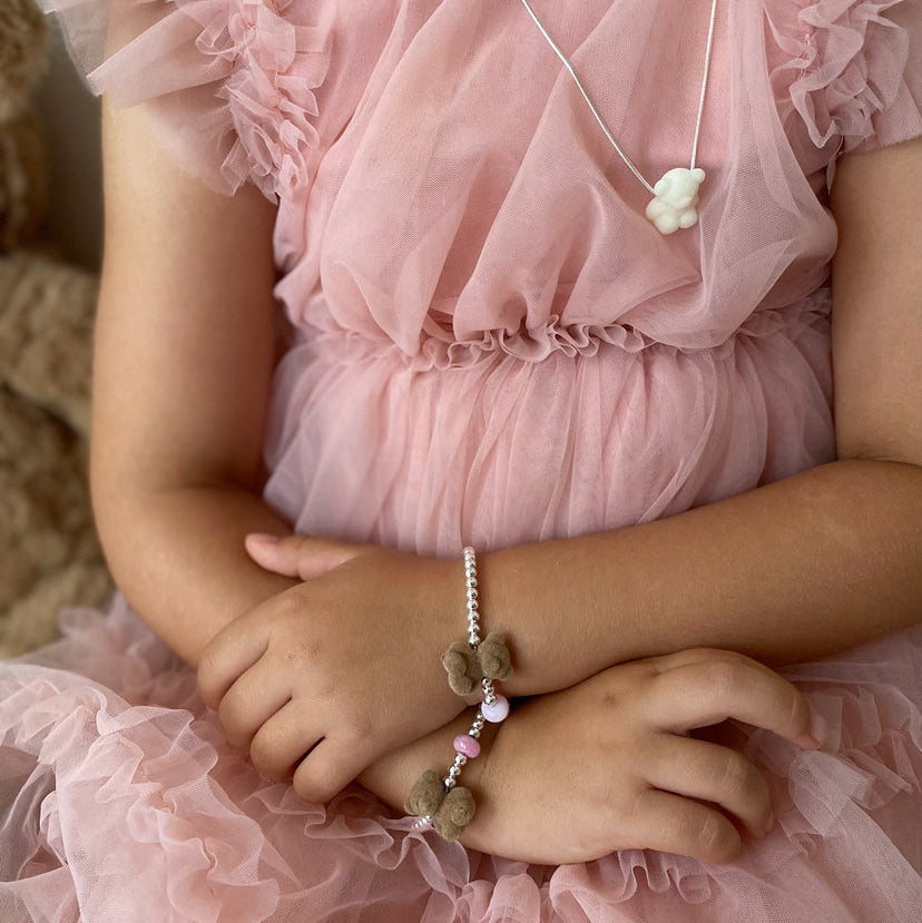 A close-up of a child wearing a pink ruffled tulle dress, a silver necklace with a white bear charm, and a beaded bracelet with brown bear pendants and pink beads.